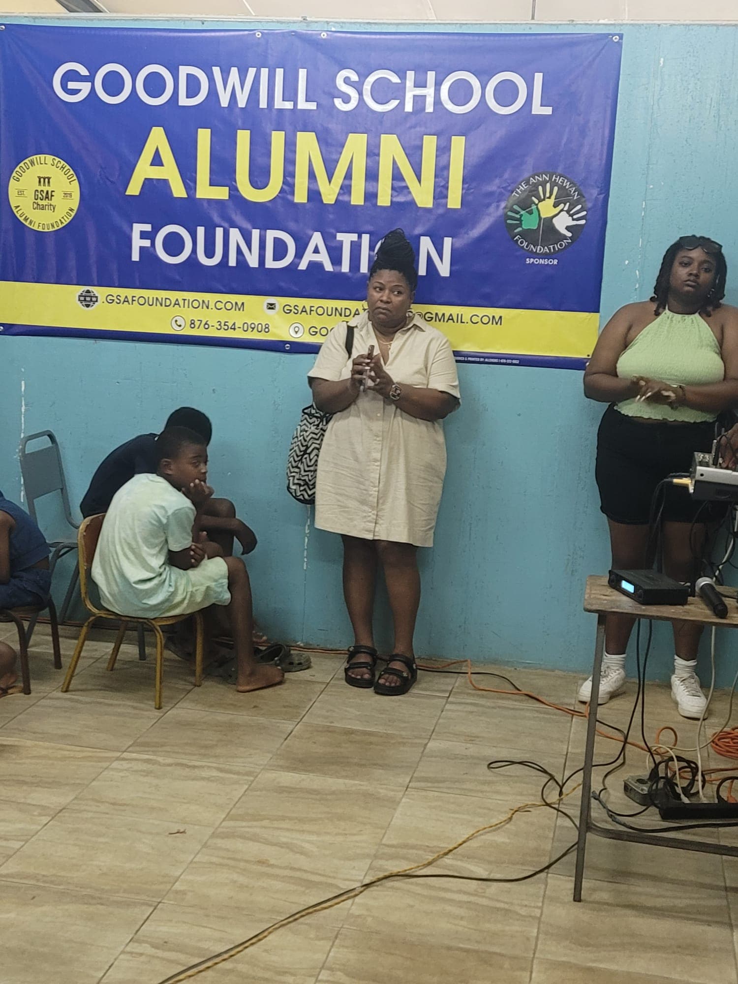 Indoor ceremony beneath a Goodwill School Alumni Foundation banner listing The Ann Hewan Foundation as a sponsor; a host addresses seated students and families
