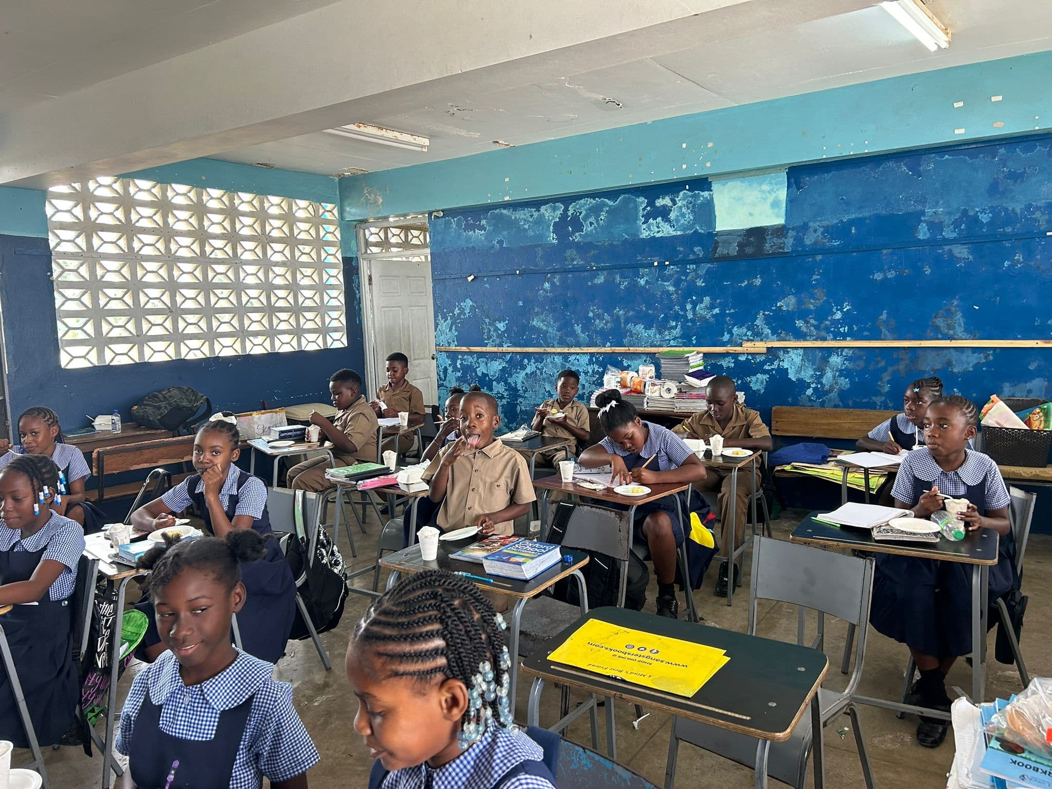Students in tan primary-school uniforms seated at desks with ice cream cups, smiling toward the camera.