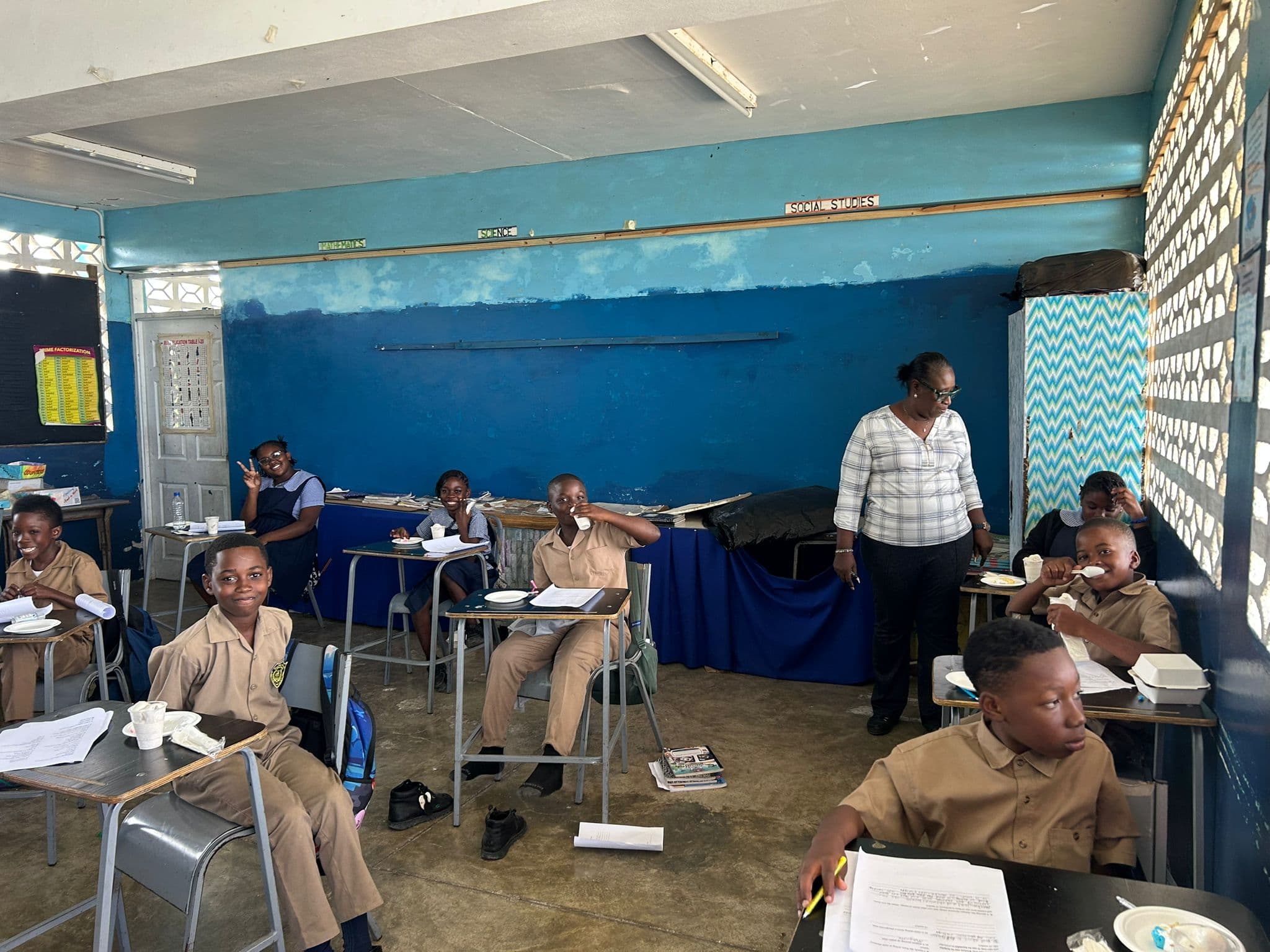 A teacher in a checked shirt stands among rows of students in tan uniforms enjoying ice cream at their desks.
