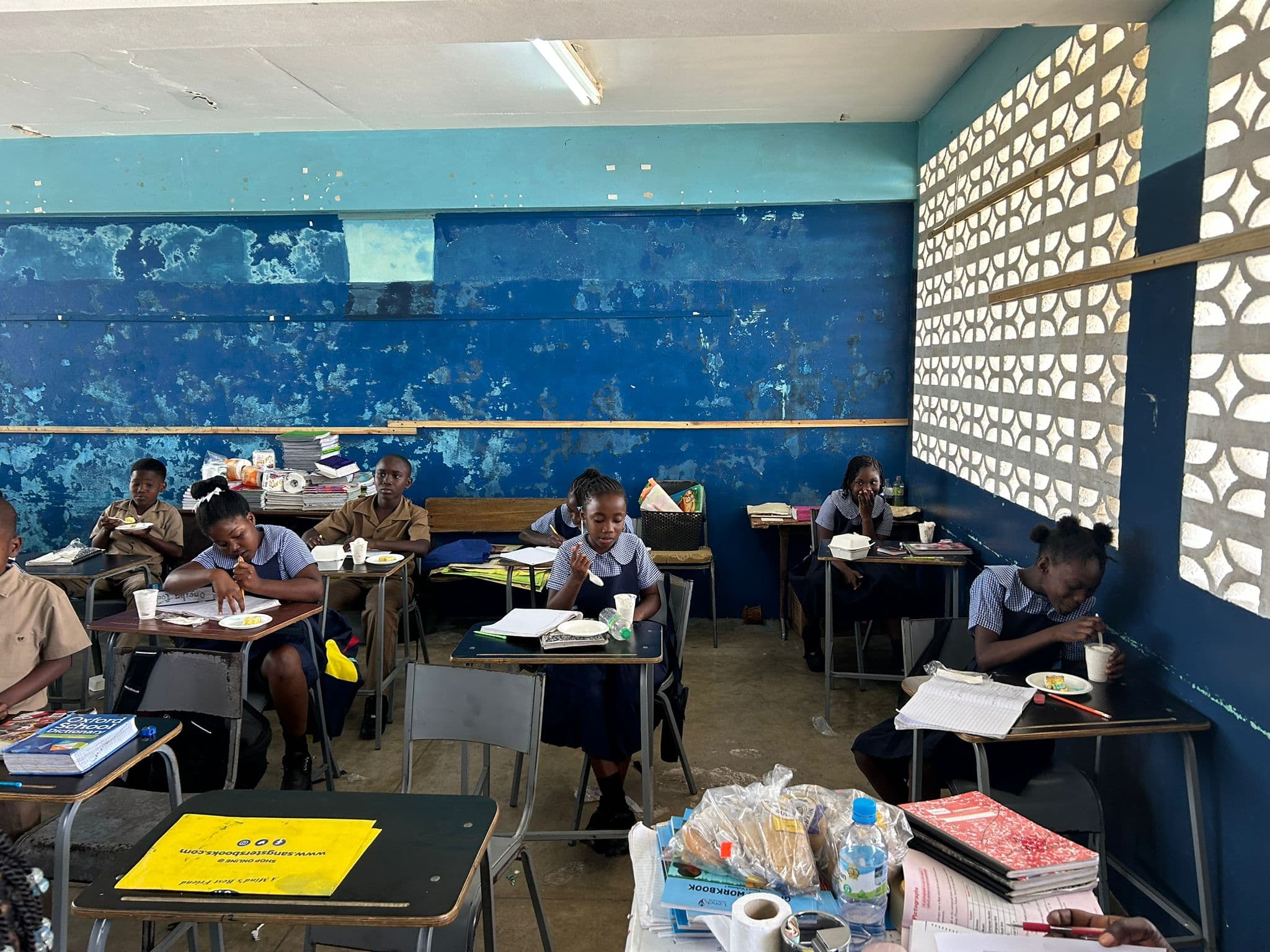Students in tan uniforms work at their desks with textbooks open and ice cream cups beside them.